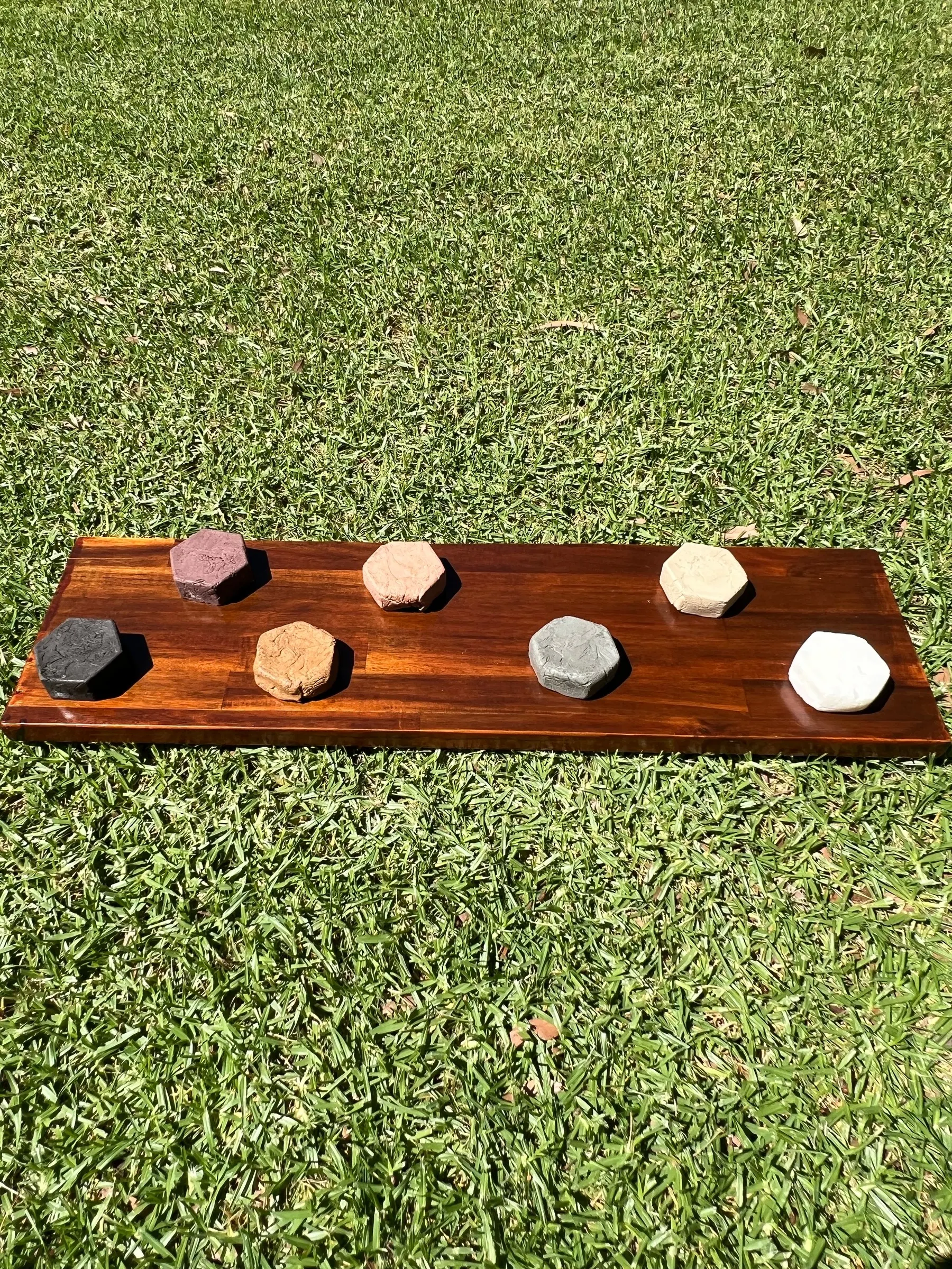 Seven hexagonal clay soaps arranged on a dark wooden board on green grass, seen from above