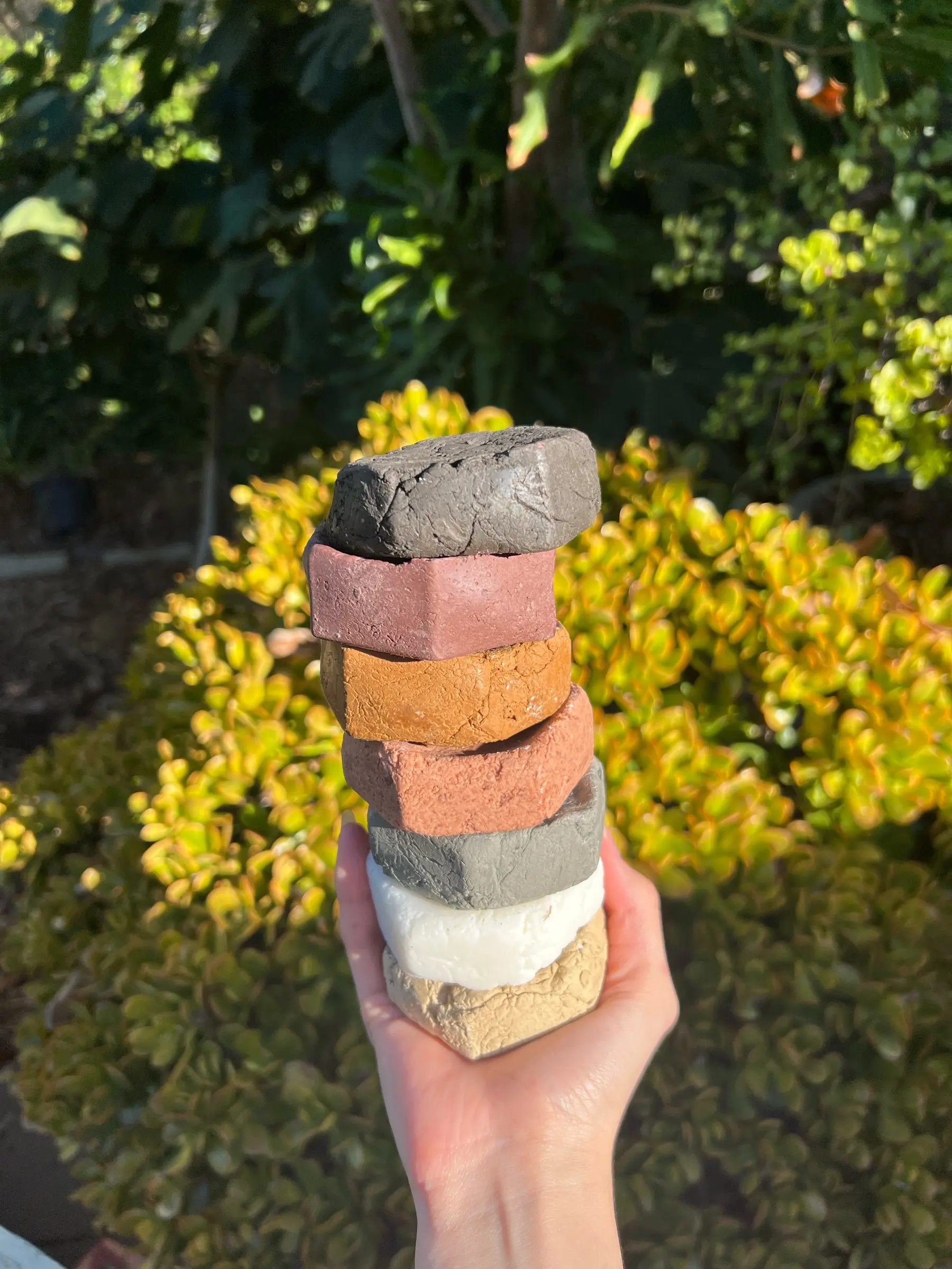 Six hexagonal clay soaps fanned out in one hand — charcoal, cream, terracotta and sand