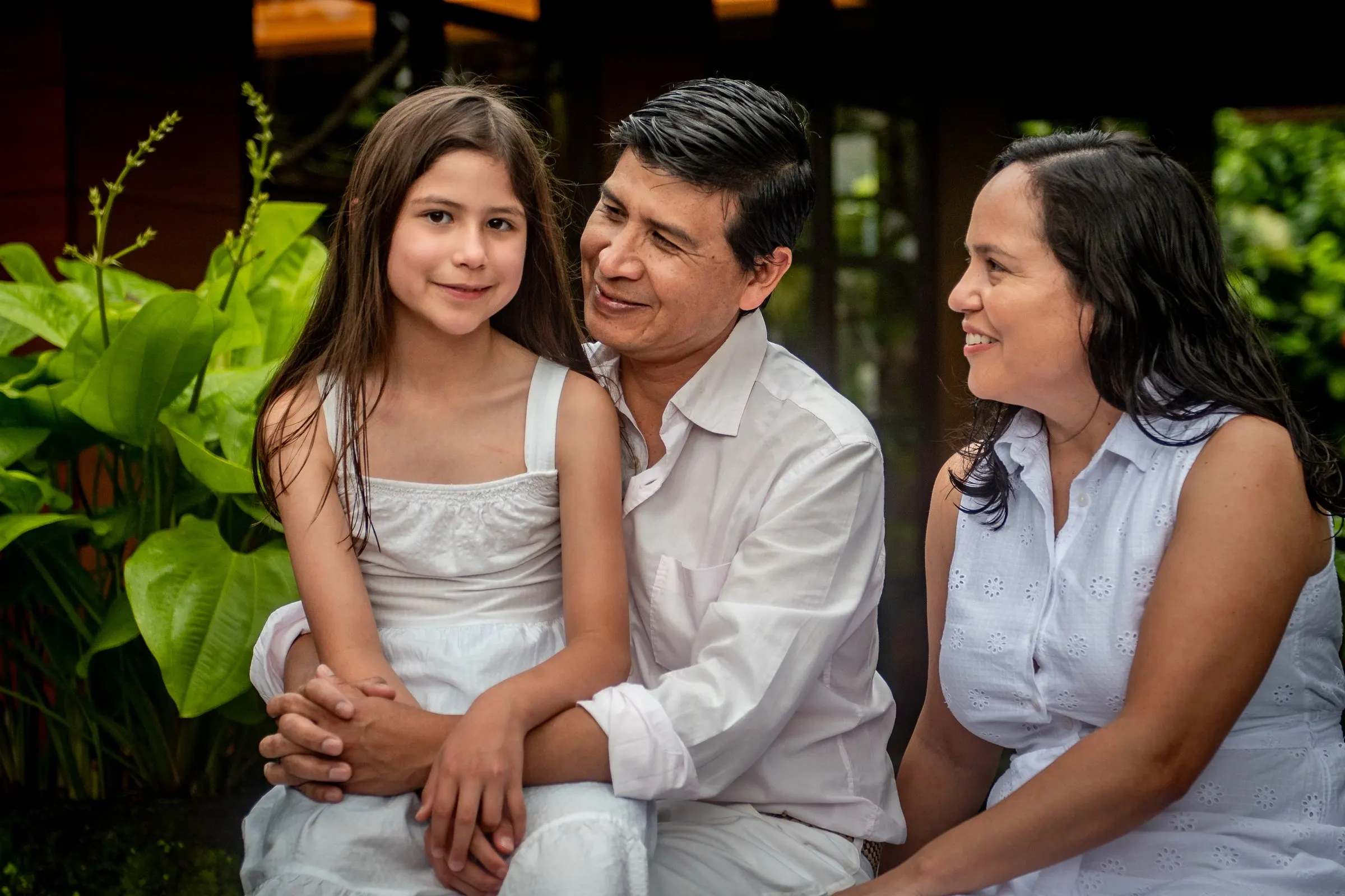 A family of three dressed in white sitting together among lush garden greenery, a young girl smiling on her father's lap