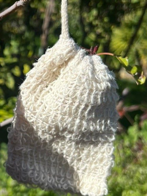 Cream cotton linen mesh soap bag with a drawstring, hanging from a branch against sunlit garden greenery